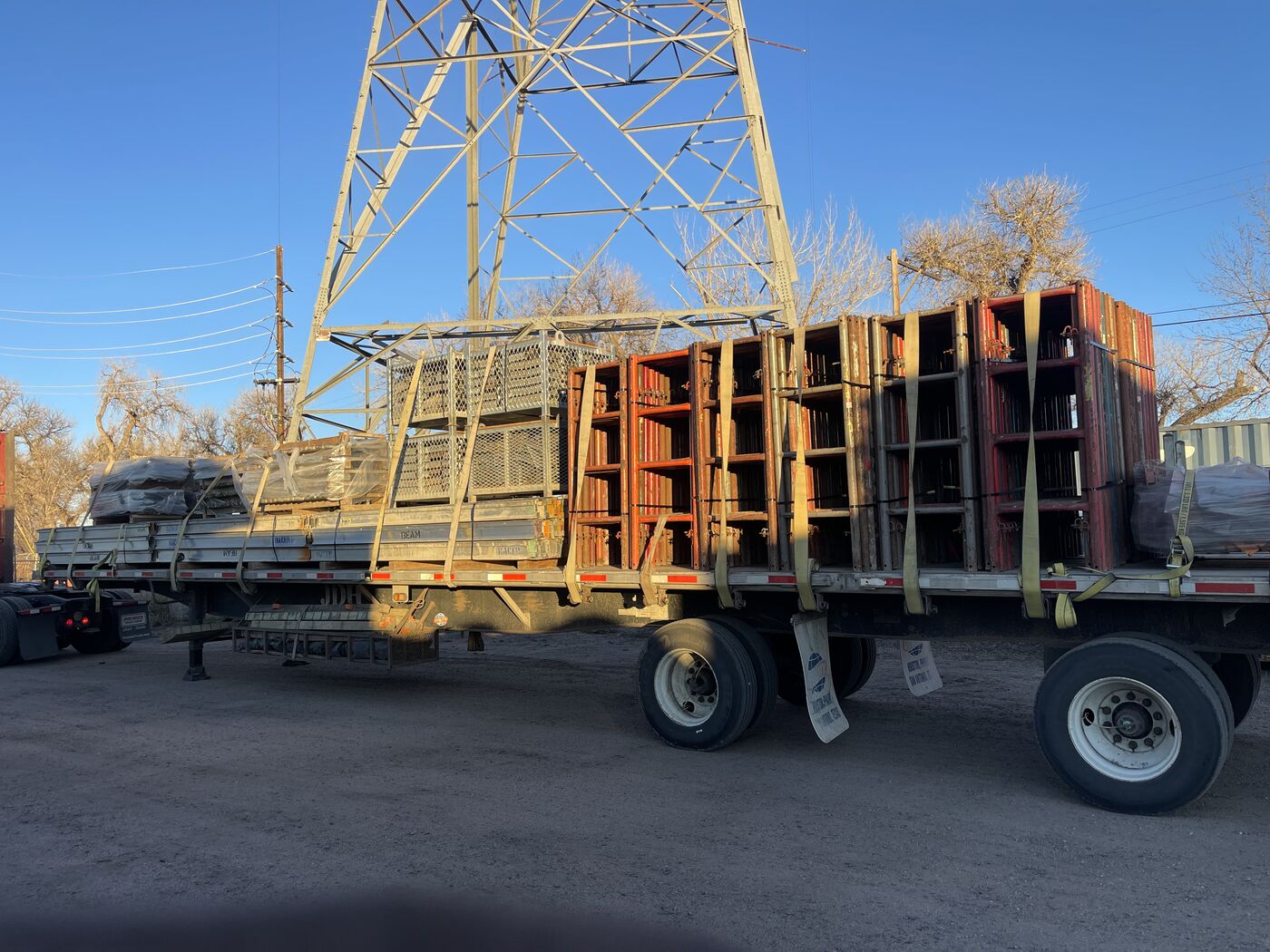 Flatbed trailer loaded with scaffolding under transmission tower at sunrise